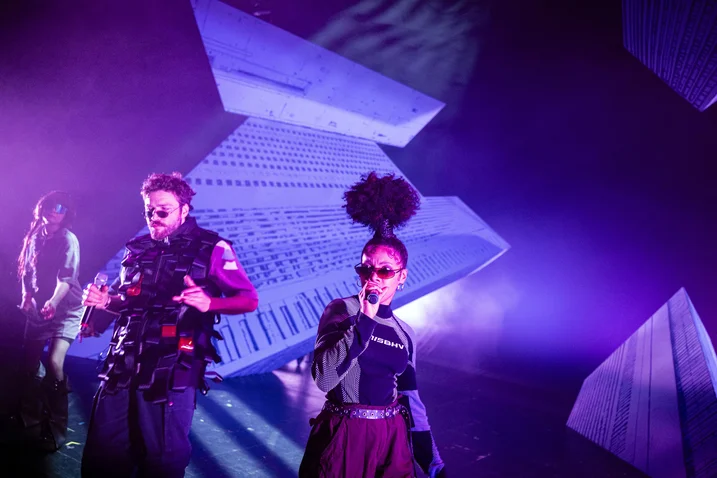 Two musicians performing on stage with futuristic decorations and purple lighting. The scene appears energetic and dynamic.