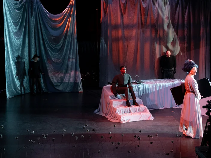 Theateraufführung auf einer Bühne mit dramatischer Beleuchtung. Ein Schauspieler sitzt, ein anderer steht, im Hintergrund sind wellenförmige Vorhänge.