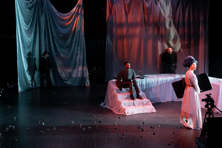 Theateraufführung auf einer Bühne mit dramatischer Beleuchtung. Ein Schauspieler sitzt, ein anderer steht, im Hintergrund sind wellenförmige Vorhänge.