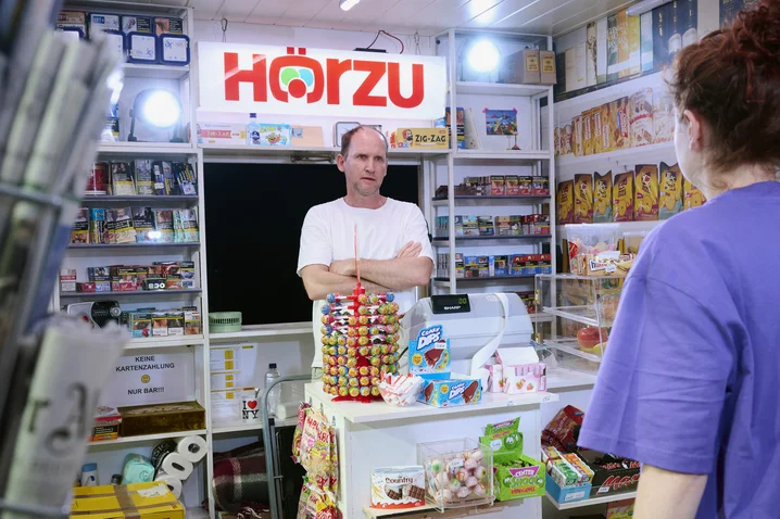 A kiosk vendor stands behind the counter, surrounded by magazines and snacks. A sign saying "Hörzu" is illuminated overhead.
