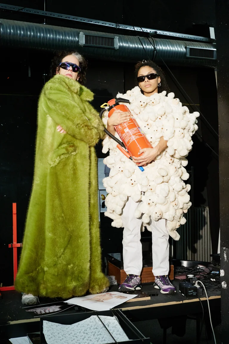 Zwei Personen, die ungewöhnliche Kleidung tragen. Eine trägt einen grünen Mantel, die andere trägt ein kunstvolles, weißes Kostüm. Beide tragen Sonnenbrillen.