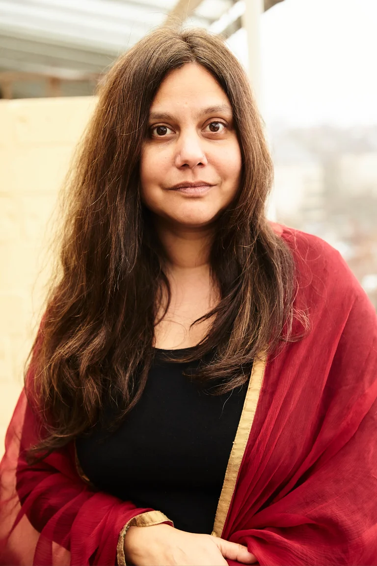 Mithu Sanyal mit langem braunem Haar, in einem schwarzen Oberteil und einem roten Schal, steht draußen. Sie schaut freundlich.