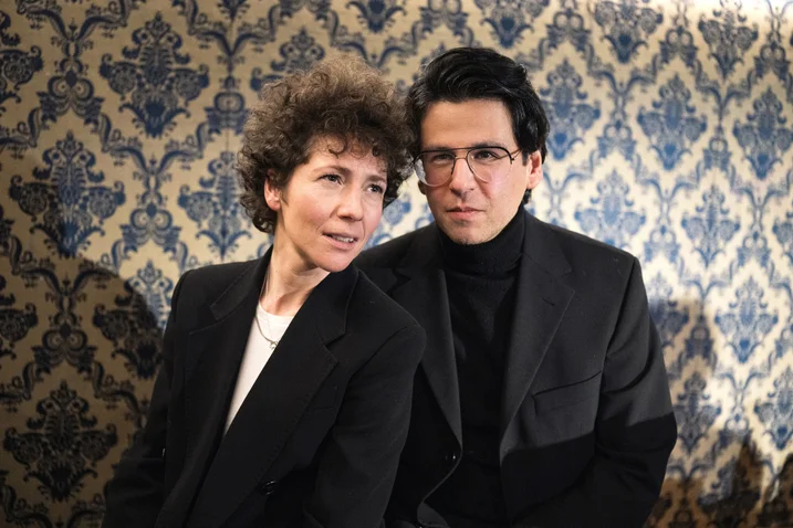 Two people in formal attire stand against a blue and gold patterned wall. Both wearing dark outfits, looking directly at the camera.