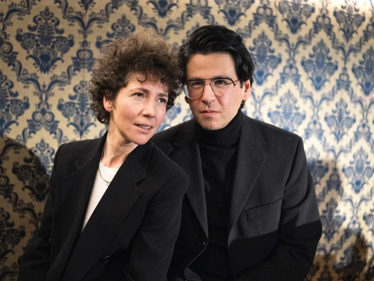 Two people in formal attire stand against a blue and gold patterned wall. Both wearing dark outfits, looking directly at the camera.