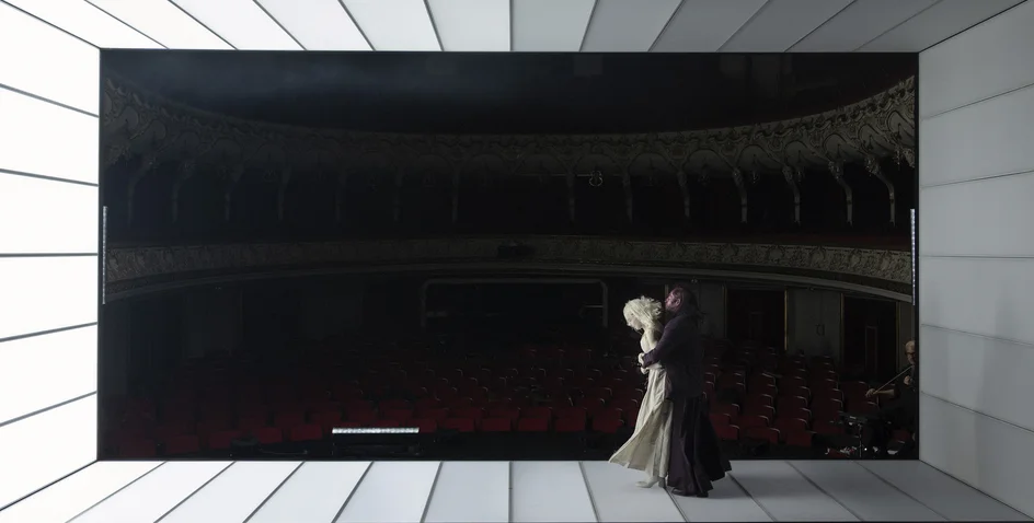 Two figures stand in a modern, minimalist space with deep darkness in the background. Their elegant attire suggests artistic context.