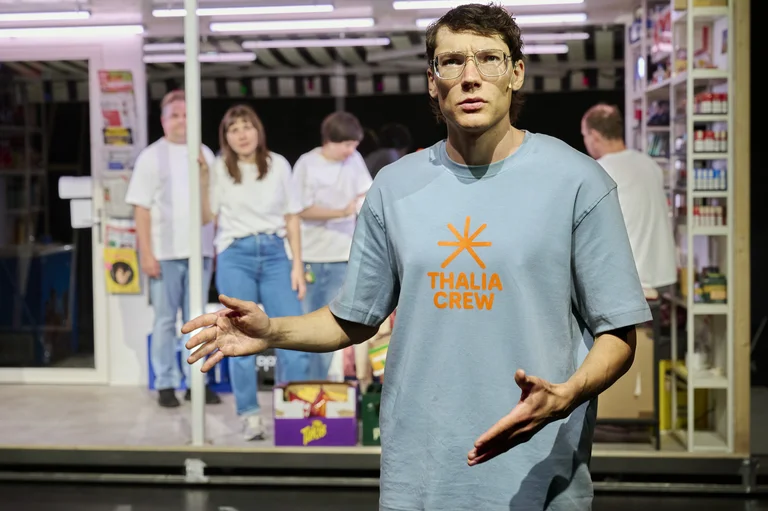 Ein Mann in einem "Thalia Crew" T-Shirt steht im Vordergrund, während andere Personen im Hintergrund in einem Laden sind.