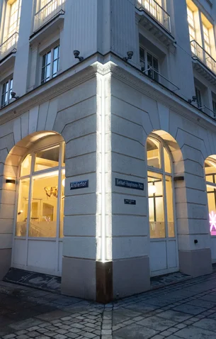 Straßenecke mit beleuchtetem Gebäude. Große Fenster geben Einblick in einen hell erleuchteten Innenraum. Straßenschilder und historische Architektur sind sichtbar.