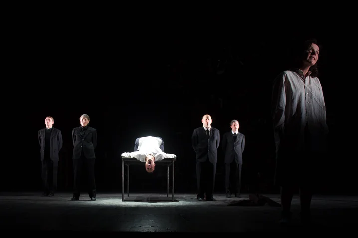 A dramatic stage scene with a lit table in the center. A person lies on it, surrounded by people in black clothing.
