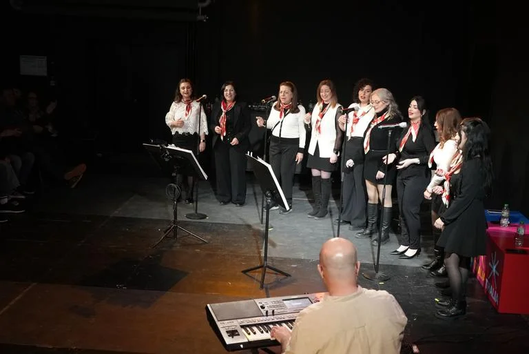 Eine Gruppe von Sängerinnen in weiß-schwarzer Kleidung steht auf einer Bühne mit Mikrofonen. Im Vordergrund ein Musiker am Keyboard.