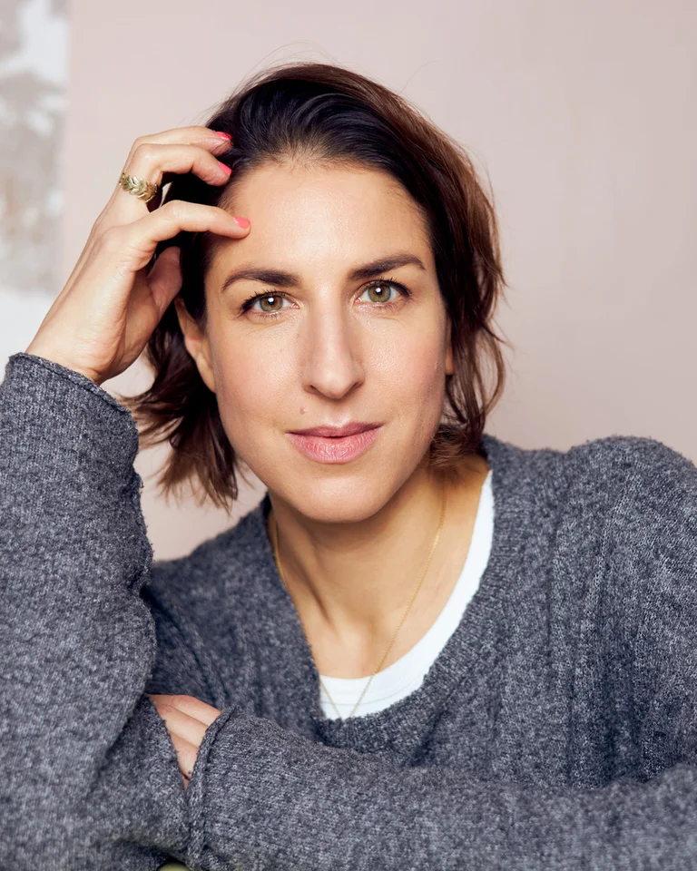 Porträtfoto von Laura de Weck. Laura de Weck ist eine Schweizer Dramatikerin und Literaturkritikerin. Auf dem Porträt ist sie mit kurzem braunem Haar und einem grauen Pullover zu sehen. Entspannt stützt sie ihren Kopf mit einer Hand ab.