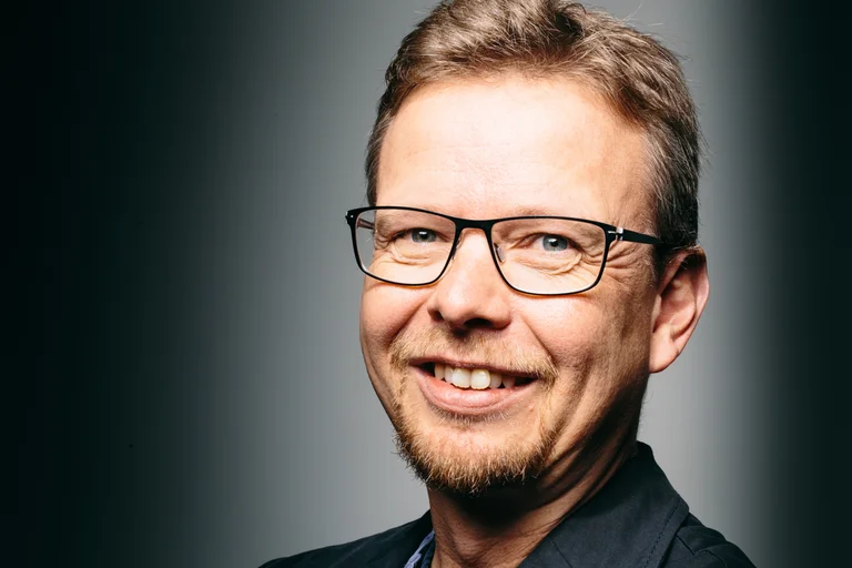 Ein Mann mit Brille und Bart lächelt freundlich. Er trägt ein dunkles Hemd. Der Hintergrund ist schlicht.