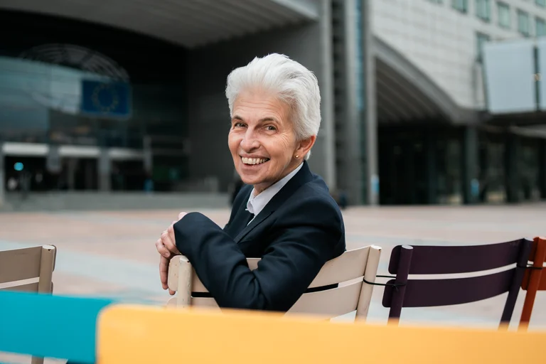 Eine lächelnde Person mit grauem Haar sitzt auf bunten Stühlen vor einem modernen Gebäude. Sie trägt einen dunklen Anzug.