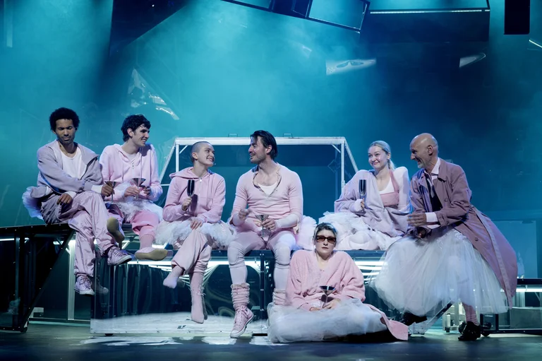 A group of actors on stage, all wearing pink outfits, sitting and chatting, under blue stage lighting.