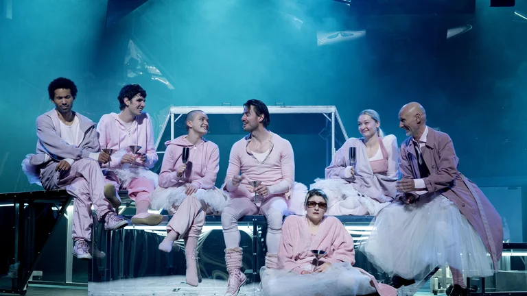 A group of actors on stage, all wearing pink outfits, sitting and chatting, under blue stage lighting.