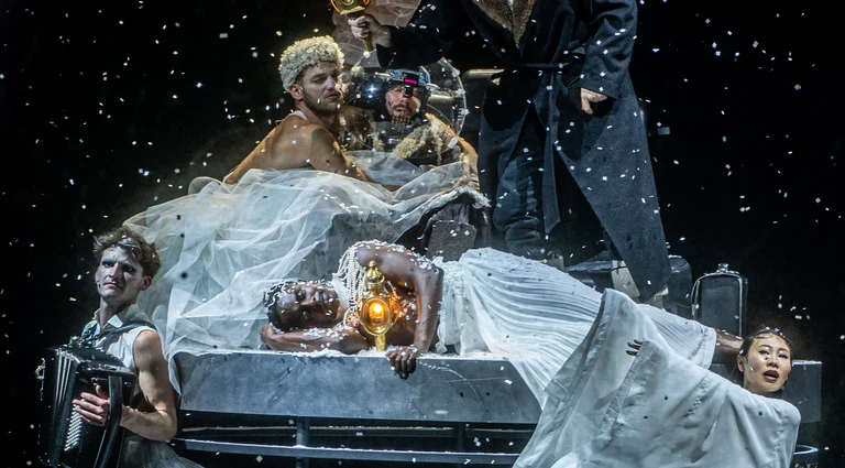 Theaterszene mit Darstellern in viktorianischer Kleidung, bedeckt mit Schnee, eine düstere und dramatische Atmosphäre erzeugend. Ein Akkordeon ist zu sehen.