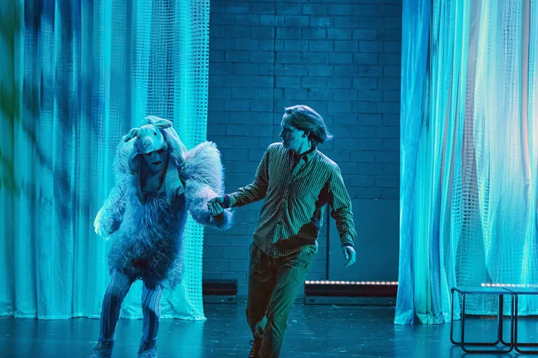 Zwei Schauspieler auf einer Bühne, einer in Vogel- und einer in menschlicher Kostümierung, tanzen in blauem Licht.