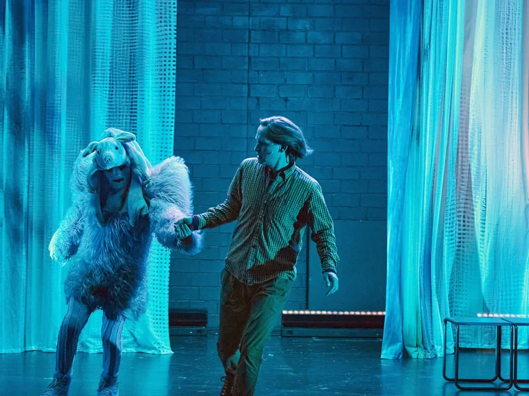 Zwei Schauspieler auf einer Bühne, einer in Vogel- und einer in menschlicher Kostümierung, tanzen in blauem Licht.