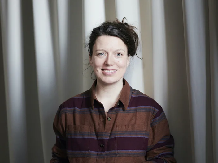 Person in a striped shirt stands smiling in front of a light-colored curtain, looking directly at the camera.