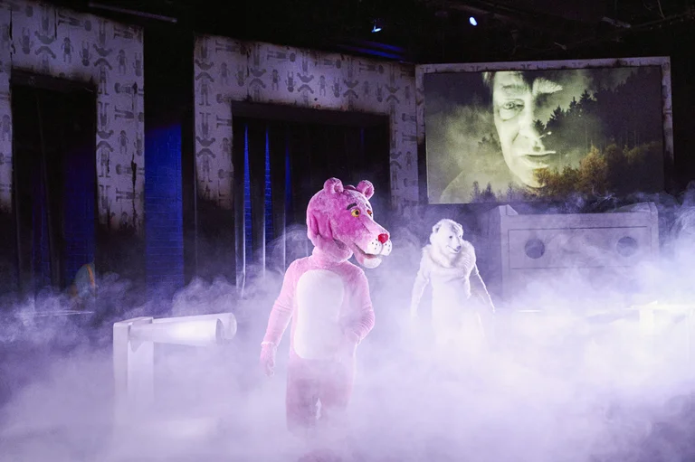 Bühnenaufführung mit einer Person im rosa Tierkostüm und einer Figur in Weiß, in Nebel gehüllt. Projektierter Hintergrund zeigt ein Gesicht.