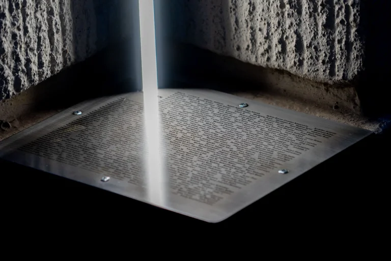 Ein Lichtstrahl fällt auf eine Metallplatte mit Inschrift, umgeben von rauen Steinmauern. Eine stimmungsvolle und nachdenkliche Szenerie.