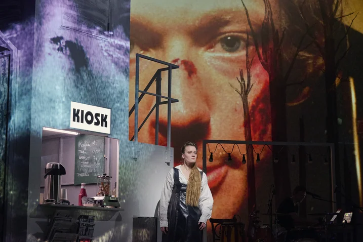 Stage set featuring a dramatic projection of a face. A performer stands in a kiosk setup with artistic, layered lighting.