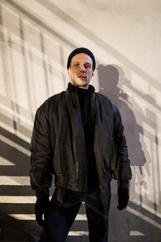 Ein Mensch steht vor einer weißen Wand, beleuchtet von seitlichem Licht, das lange Schatten wirft. Sie tragen eine dunkle Jacke und Mütze.