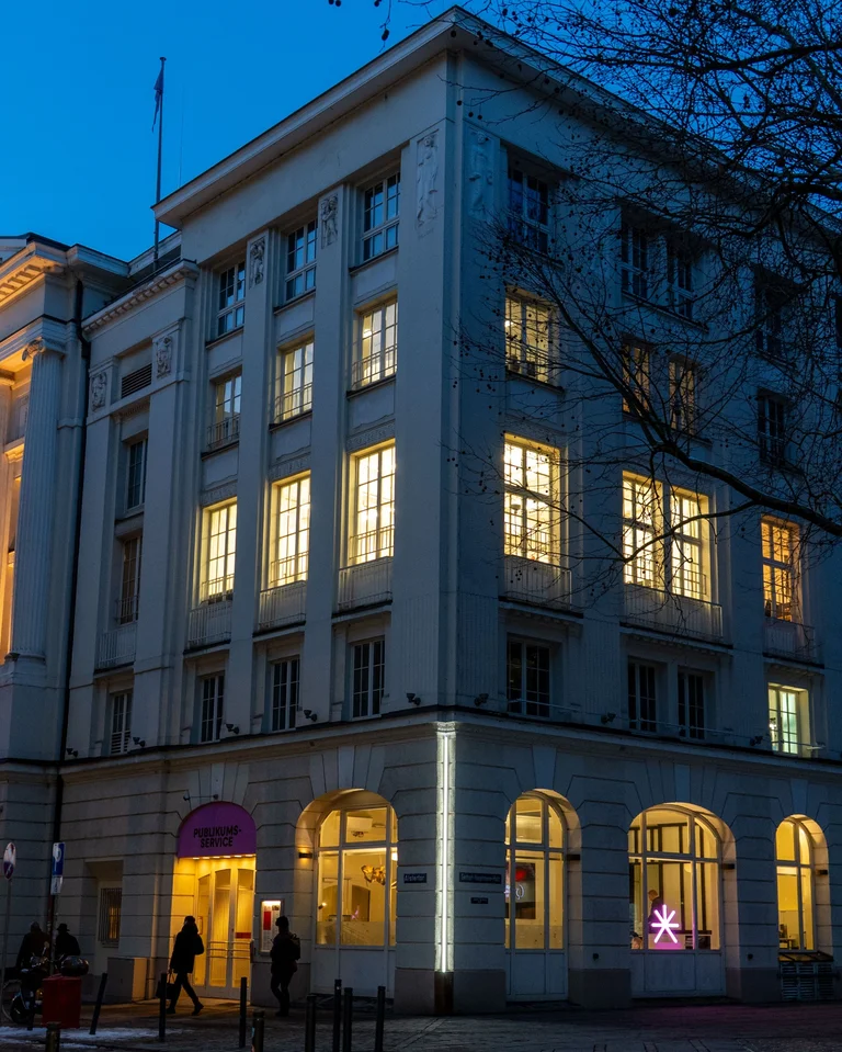Ein beleuchtetes Gebäude bei Nacht, mit mehreren Fenstern. Fußgänger spazieren an der Straße entlang. Einige Fenster leuchten warm.