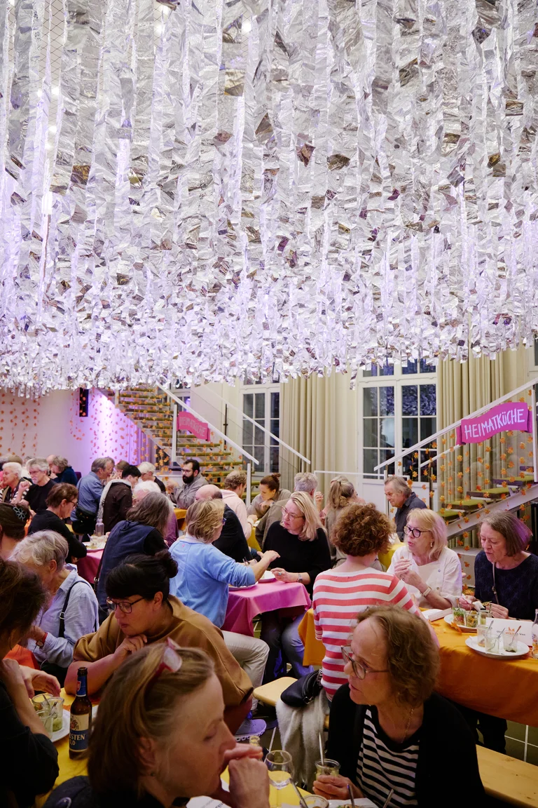 Ein geschäftiger Raum voller Menschen, die an Tischen unter einer kunstvollen, funkelnden Deckeninstallation sitzen. Lebendige und einladende Atmosphäre.