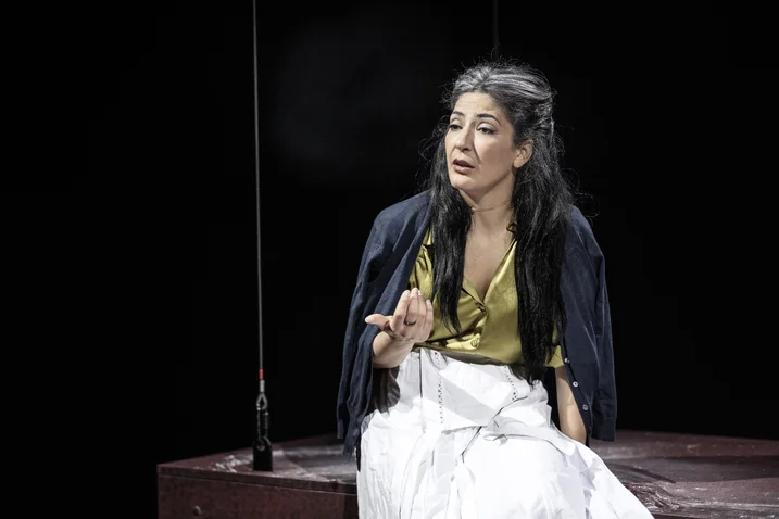 A woman with long hair sits on stage. She wears a yellow top and white skirt, gesturing expressively.