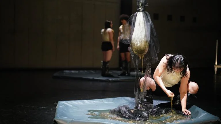Performance-Kunst auf einer Bühne, zwei Personen interagieren mit Wasser in einem künstlerischen Ausdruck. Dunkle, minimale Beleuchtung.