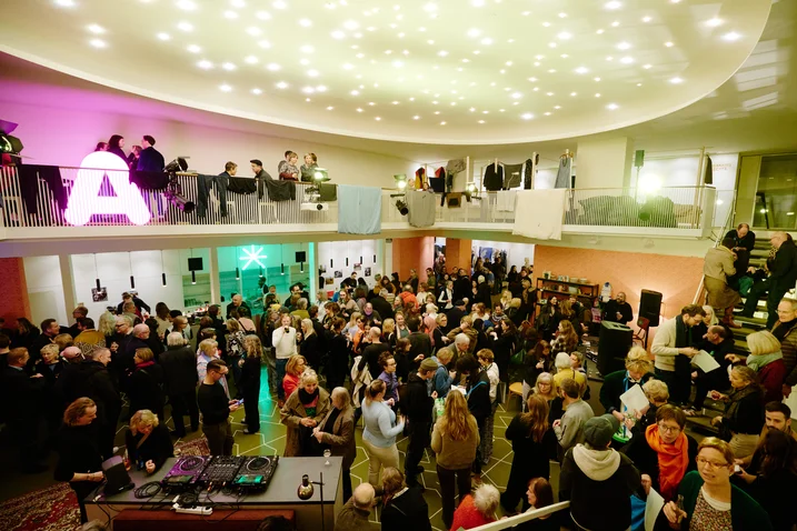 A large crowd in a stylish event space with modern lighting. Many are socializing, some on an upper gallery level.