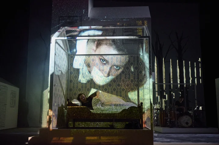 A stage scene with a projected, oversized close-up of a face over a reclining actor within a modern set design.
