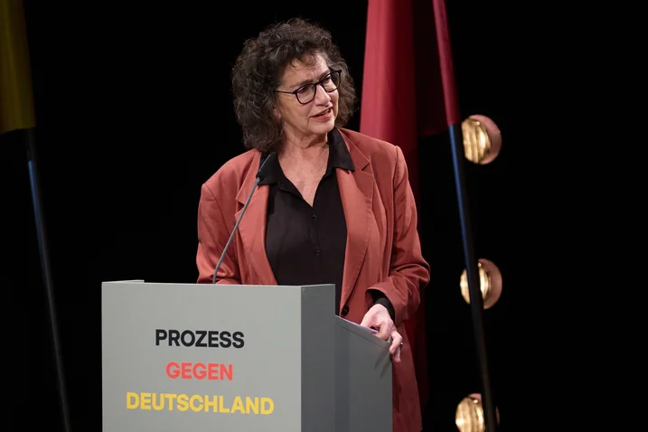 Eine Frau spricht bei einer Veranstaltung mit dem Thema "Prozess gegen Deutschland". Sie steht hinter einem Rednerpult.