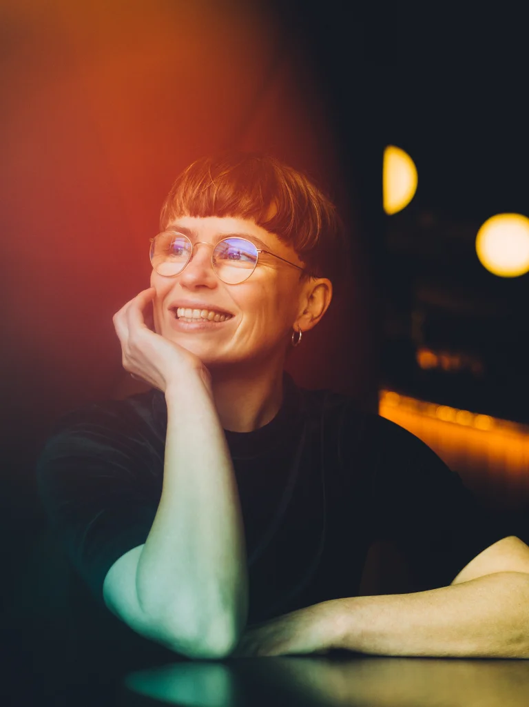 Caren Jeß mit Brille lächelt verträumt, gestützt auf die Hand, umgeben von warmem Licht und dunklem Hintergrund.