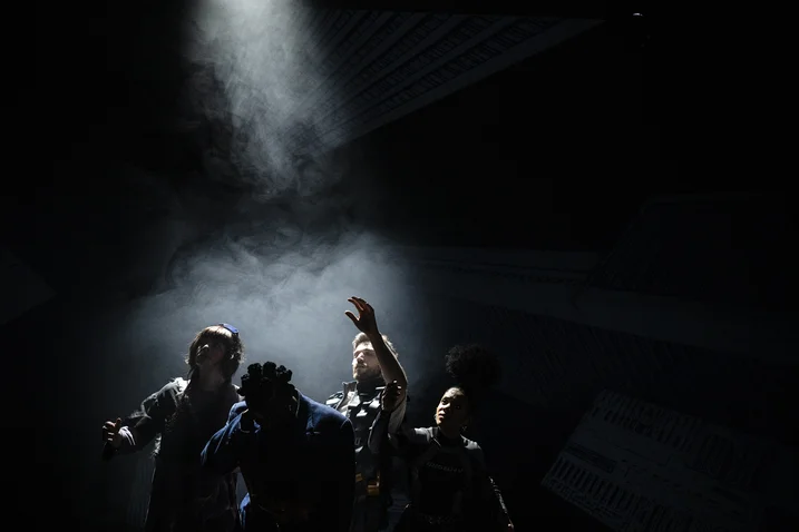 Dark stage scene with four people under a single beam of light. They appear to be standing in intense concentration.
