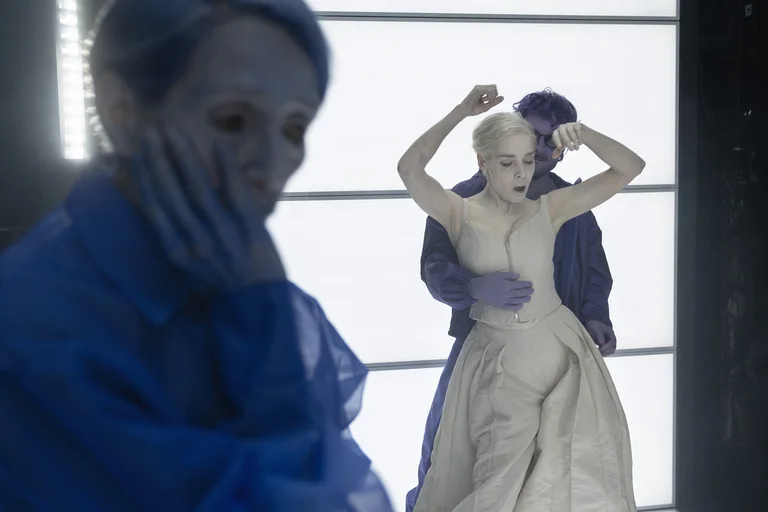 Three individuals in a theatrical setting. One in the foreground holds their hand to their face, as a couple dances behind.