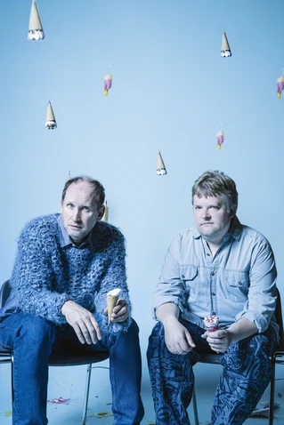 Two men sit on chairs against a blue background. Ice cream cones hang from the ceiling, creating a whimsical atmosphere.