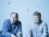 Two men sit on chairs against a blue background. Ice cream cones hang from the ceiling, creating a whimsical atmosphere.