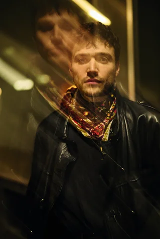 Ein Mann in einer Lederjacke mit einem bunten Schal, künstlerisch doppelt belichtet, steht in einem dunklen, beleuchteten Innenraum.