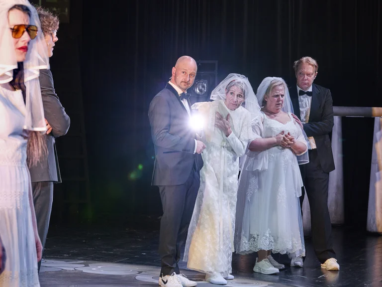 Eine Bühnenszene zeigt Leute in Hochzeitskleidung, einige tragen Brillen. Ein Mann im Anzug hält ein helles Licht.