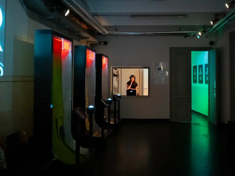 Ein dunkler Raum mit erleuchteten Kabinen und einem hell erleuchteten Fenster, in dem eine Person sichtbar ist.