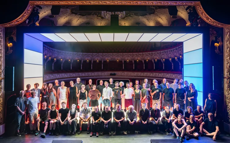 Ein großes Gruppenfoto auf einer Theaterbühne, in einem prächtig dekorierten klassischen Theater. Viele Menschen, vorne sitzend und dahinter stehend.