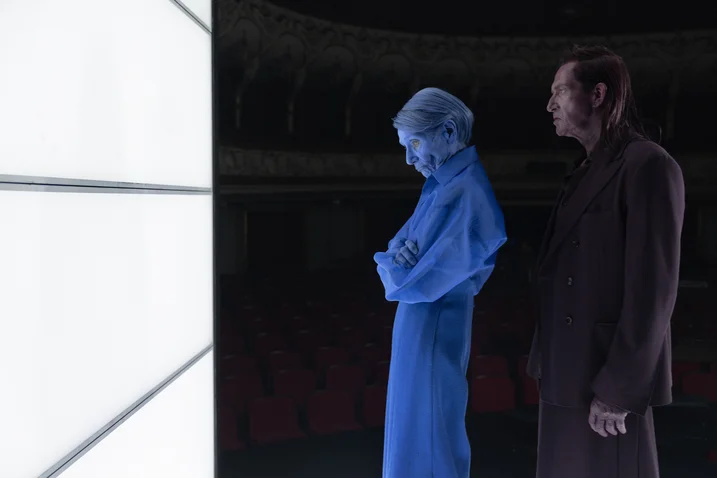 Two figures stand in front of a glowing screen. One appears blue and translucent, the other wears a dark coat.