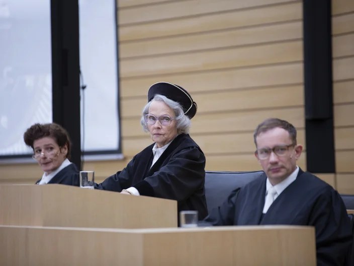 Drei Richter in schwarzer Robe sitzen auf einer erhöhten Plattform in einem Gerichtssaal. Die mittlere Richterin trägt eine Richterkappe.