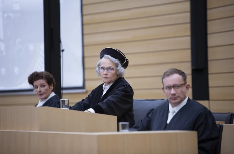 Drei Richter in schwarzer Robe sitzen auf einer erhöhten Plattform in einem Gerichtssaal. Die mittlere Richterin trägt eine Richterkappe.