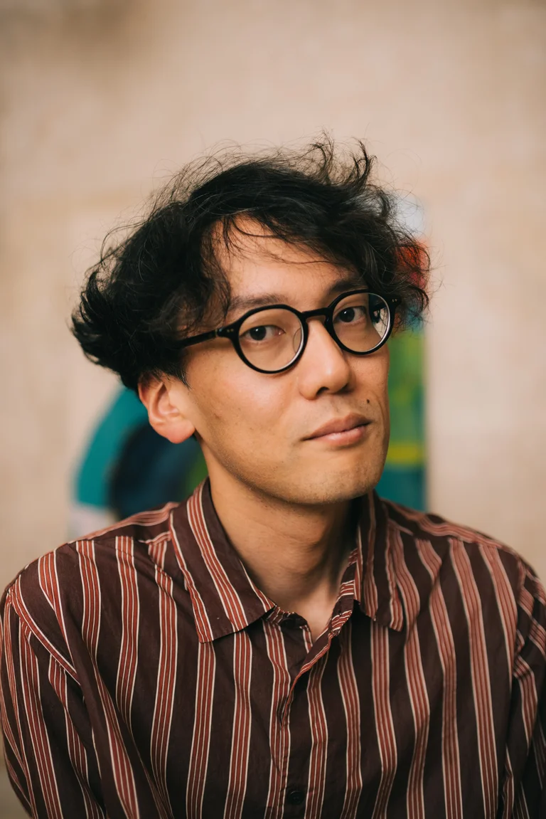 Ein Mann mit lockigem Haar und Brille trägt ein gestreiftes Hemd. Der Hintergrund ist unscharf mit bunten Farben.