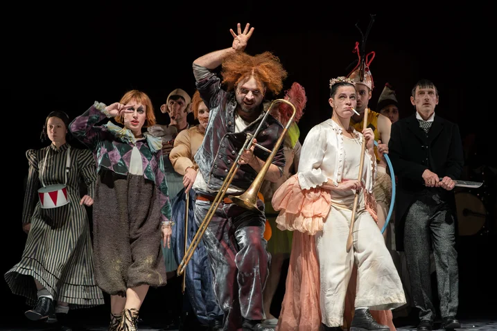 A group of performers in colorful, eccentric costumes on stage, with one playing a trombone and others gesturing animatedly.
