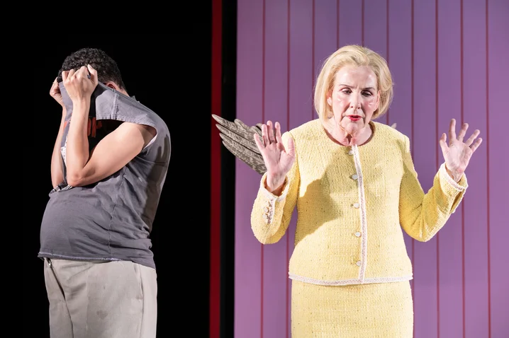 Two people stand on stage; one covers their face with hands, the other in yellow raises their hands.