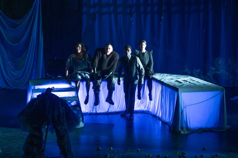 Vier Personen auf einer Bühne, beleuchtet von blauem Licht. Sie sitzen auf einer erhöhten Plattform in einer dunklen Theaterkulisse.