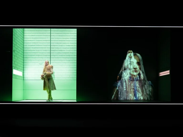 Zwei Personen auf einer Bühne; links in hellem Licht, rechts in schillerndem Gewand. Die Szene wirkt futuristisch und kunstvoll.